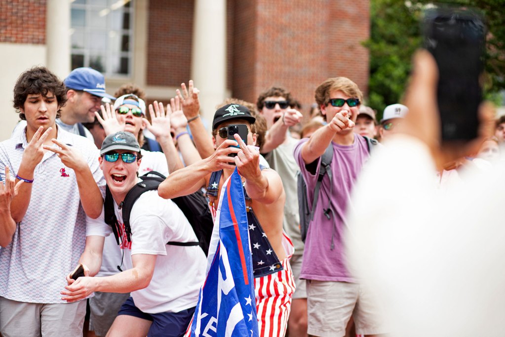 a photo shows white male ole miss students pointing, flipping the bird, laguhing and holding a trump flag